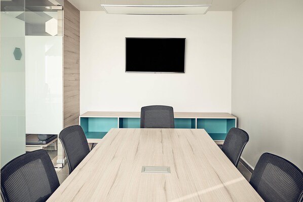 A conference room with a long table, four chairs, a flat-screen TV on the wall, and a glass partition on one side.