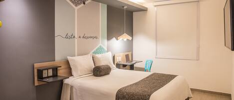 A modern hotel room with a bed, bedside table, and a desk with a chair.