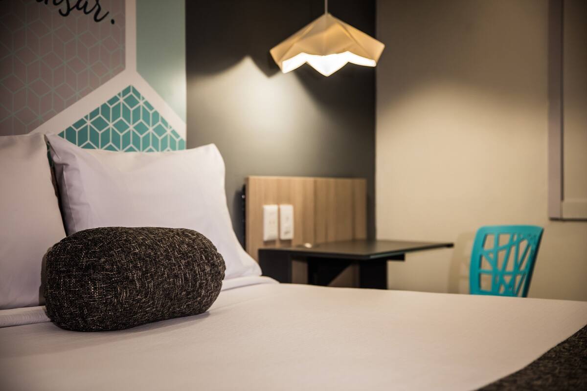 Un lit bien fait, agrémenté d’un coussin texturé, une table en bois et une chaise bleue dans une pièce moderne aux murs ornés de motifs géométriques et dotée d’un luminaire suspendu.