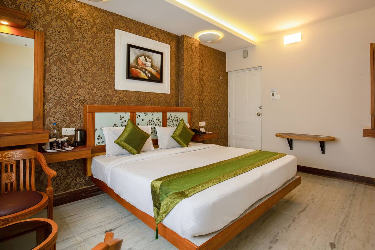 Quarto de hotel com uma cama grande, mesa de cabeceira de madeira, uma cadeira, um espelho e um quadro na parede.