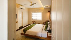 A hotel room with a bed, a ceiling fan, an air conditioning unit, a window with blinds, a chair, a bedside table, and a telephone. - Treebo The Qasr (Kanayannur)