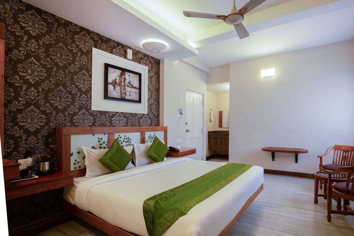 Quarto com cama, mesa de cabeceira, cadeira, ventoinha no teto e banheiro visível por uma porta aberta.