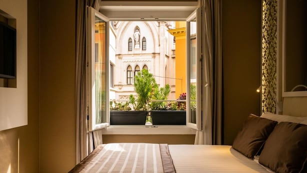 Quarto de hotel com uma cama grande, vista para um edifício com uma estátua e uma varanda com plantas.
