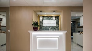 Uma área de recepção com parede bege, balcão de recepção branco e uma placa com a inscrição "RESIDENZA VENTI SETTEMBRE".