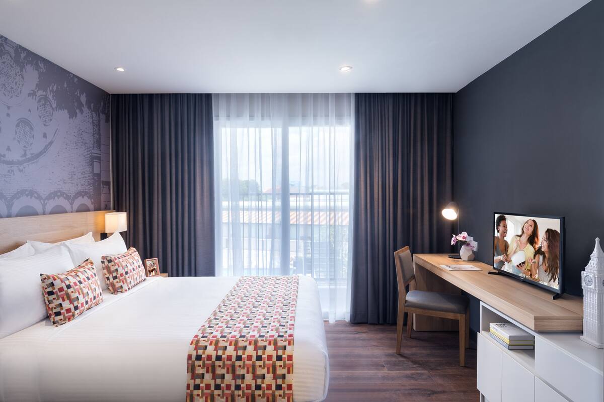 A modern hotel room with a large bed, a desk with a computer, and a view of the city through large windows.