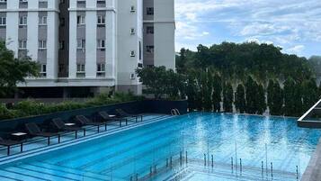 A swimming pool with lounge chairs, surrounded by a building with multiple floors and a forested area in the background.
