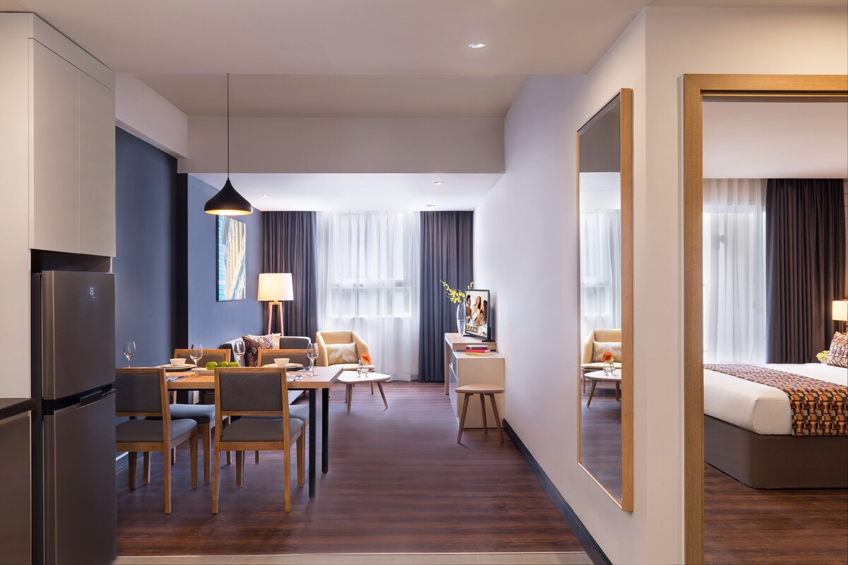 A modern hotel room with a bed, a dining area with chairs and a table, a kitchen area with a refrigerator, and a large mirror.