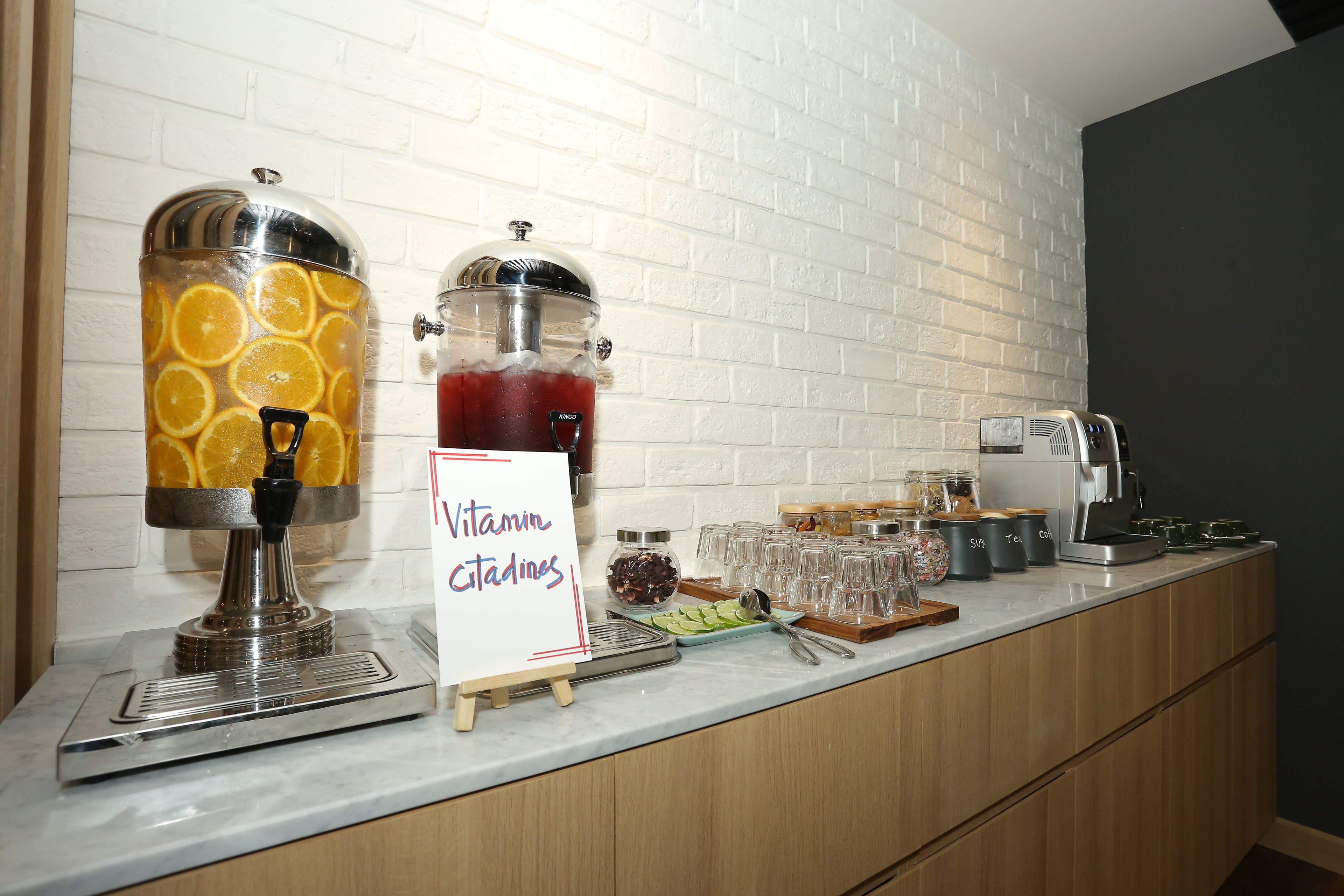 Ein Saftspender mit Orangenscheiben, ein Schild mit der Aufschrift 'Vitamin Citadines' und eine Kaffeemaschine auf einer Küchenarbeitsplatte.