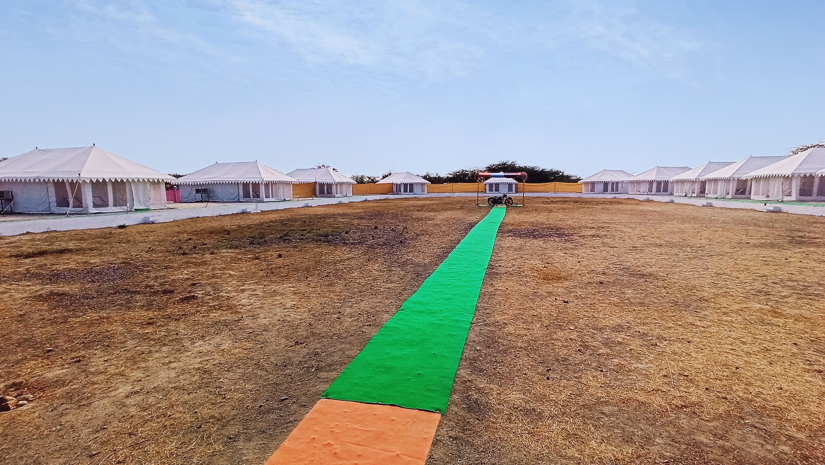 Um camping com barracas dispostas em fileiras, um caminho verde e um campo de terra.