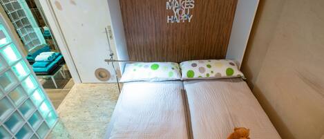 Una cama pequeña y bien hecha con un osito de peluche encima, una pared de madera con una frase motivadora, y una parte de un baño con cabina de ducha de cristal.