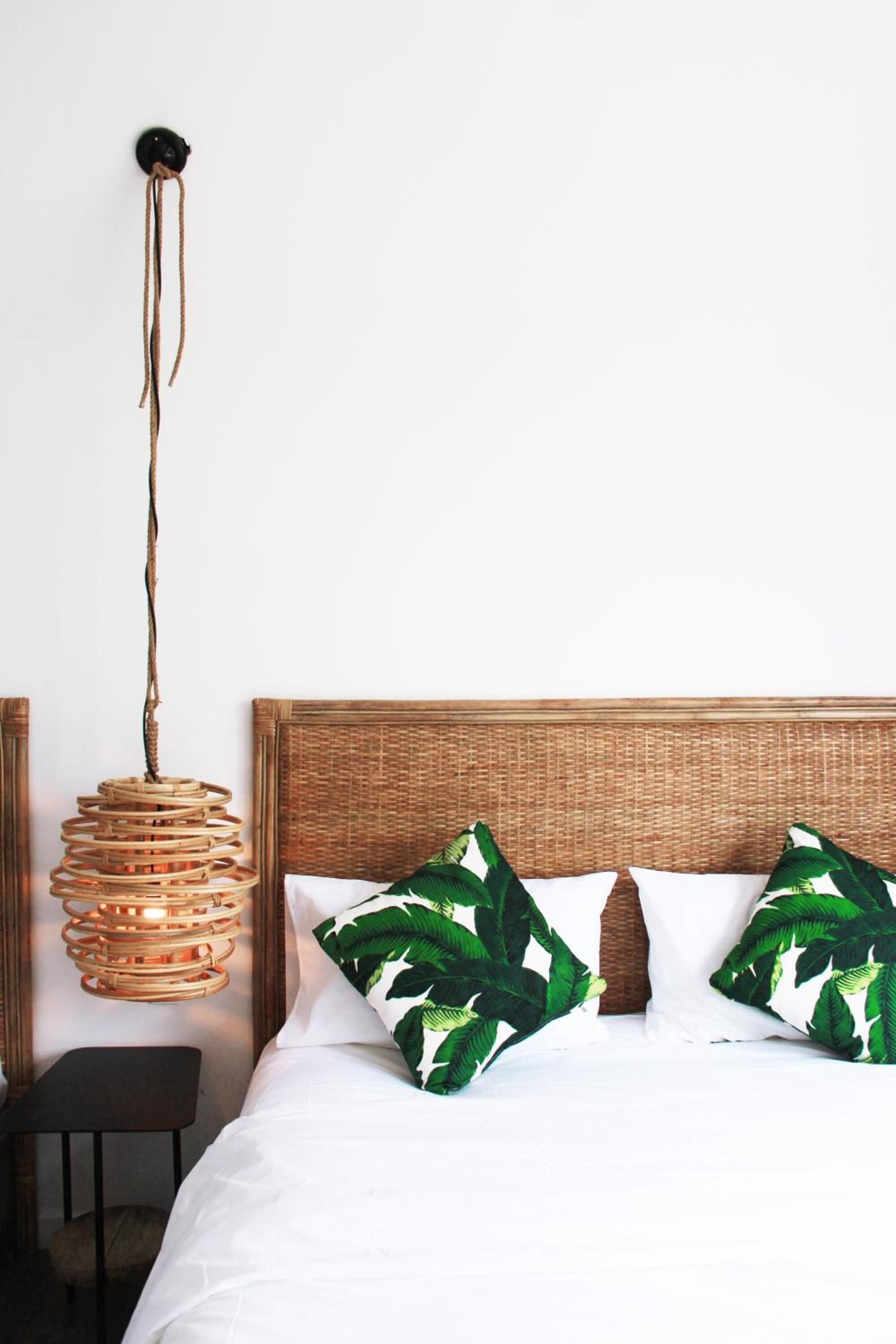 A bed with white bedding and two green leaf-patterned pillows, a woven headboard, and a hanging light fixture.