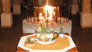 Une table ornée d’une théière à motifs floraux et d’une petite plante en pot, le tout placé devant une cheminée où l’on voit des flammes.