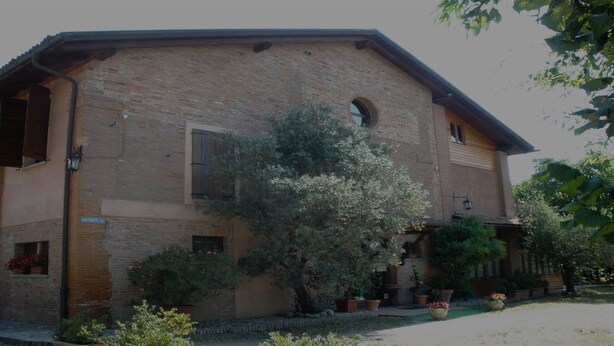 Un bâtiment rustique en briques, avec un grand arbre devant, une enseigne indiquant « ENTRATA » et des plantes en pot tout autour.