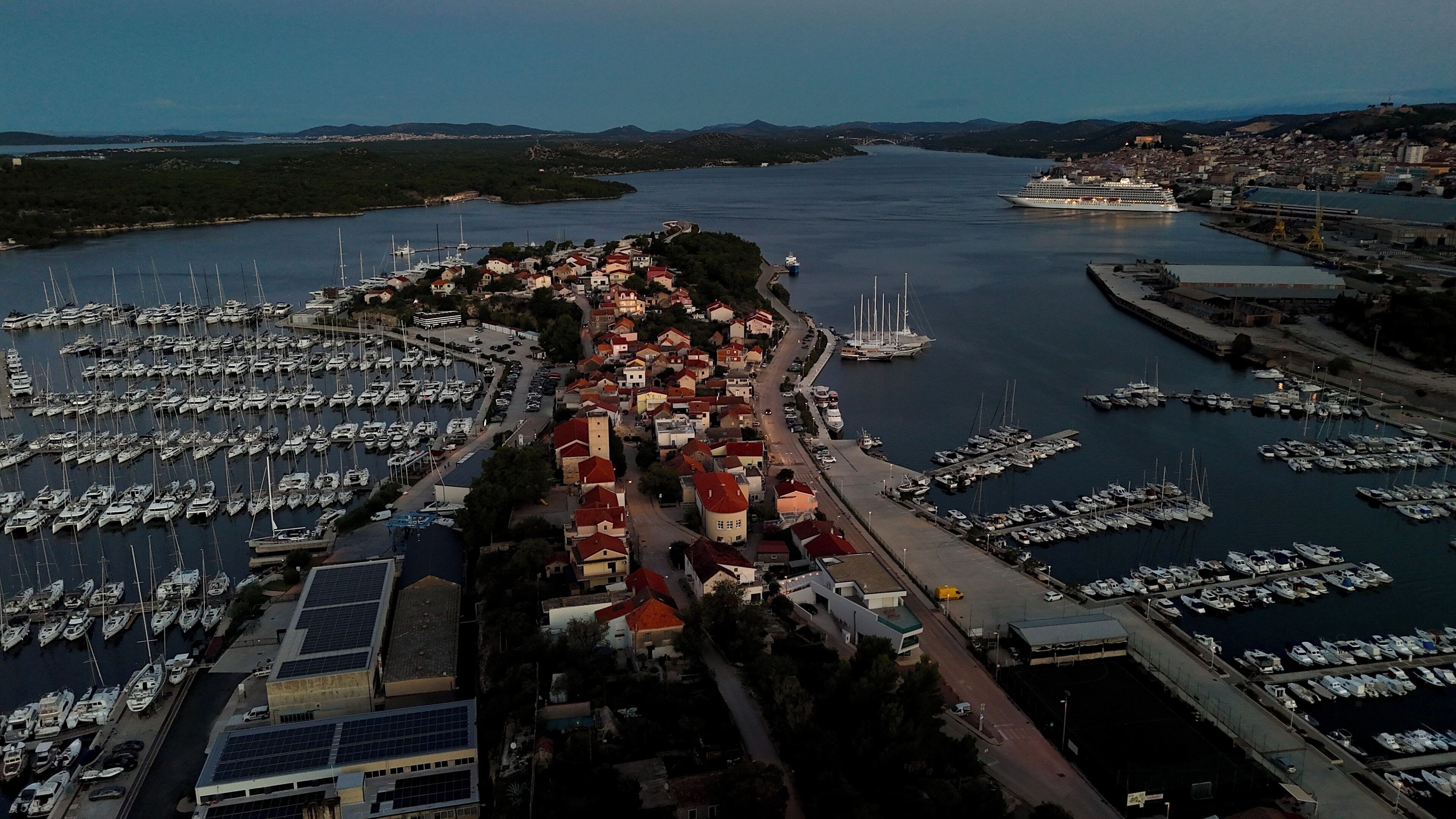 Vista aèria d'una marina amb nombrosos vaixells amarrats, una vila amb edificis de teulades vermelles i un gran vaixell de creuer al fons.