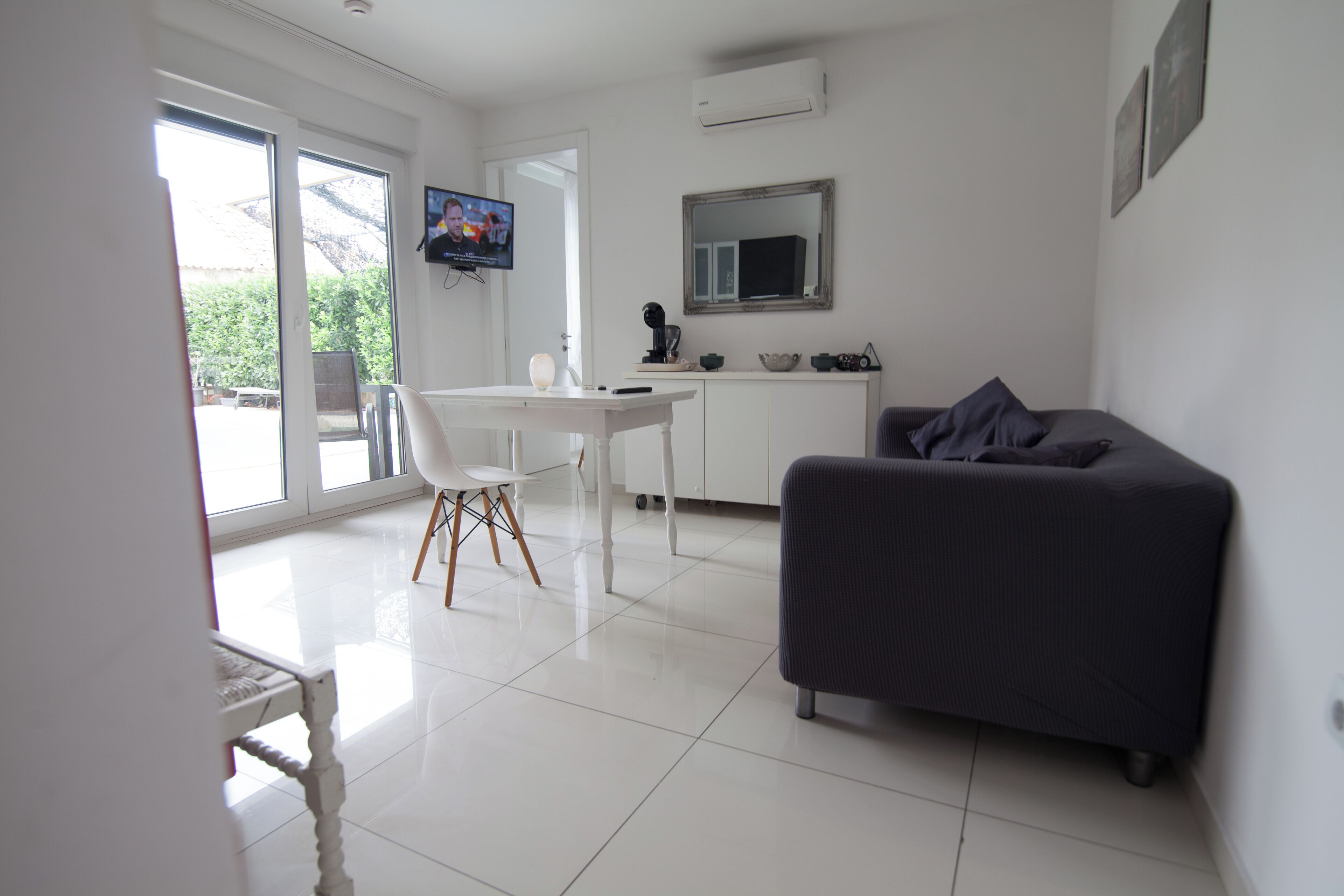 Una sala de estar moderna con un sofá oscuro, una mesa de comedor blanca y un televisor montado en la pared.