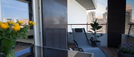 Un balcón moderno con puerta de cristal, una silla de descanso, una planta en maceta y vistas de la ciudad.
