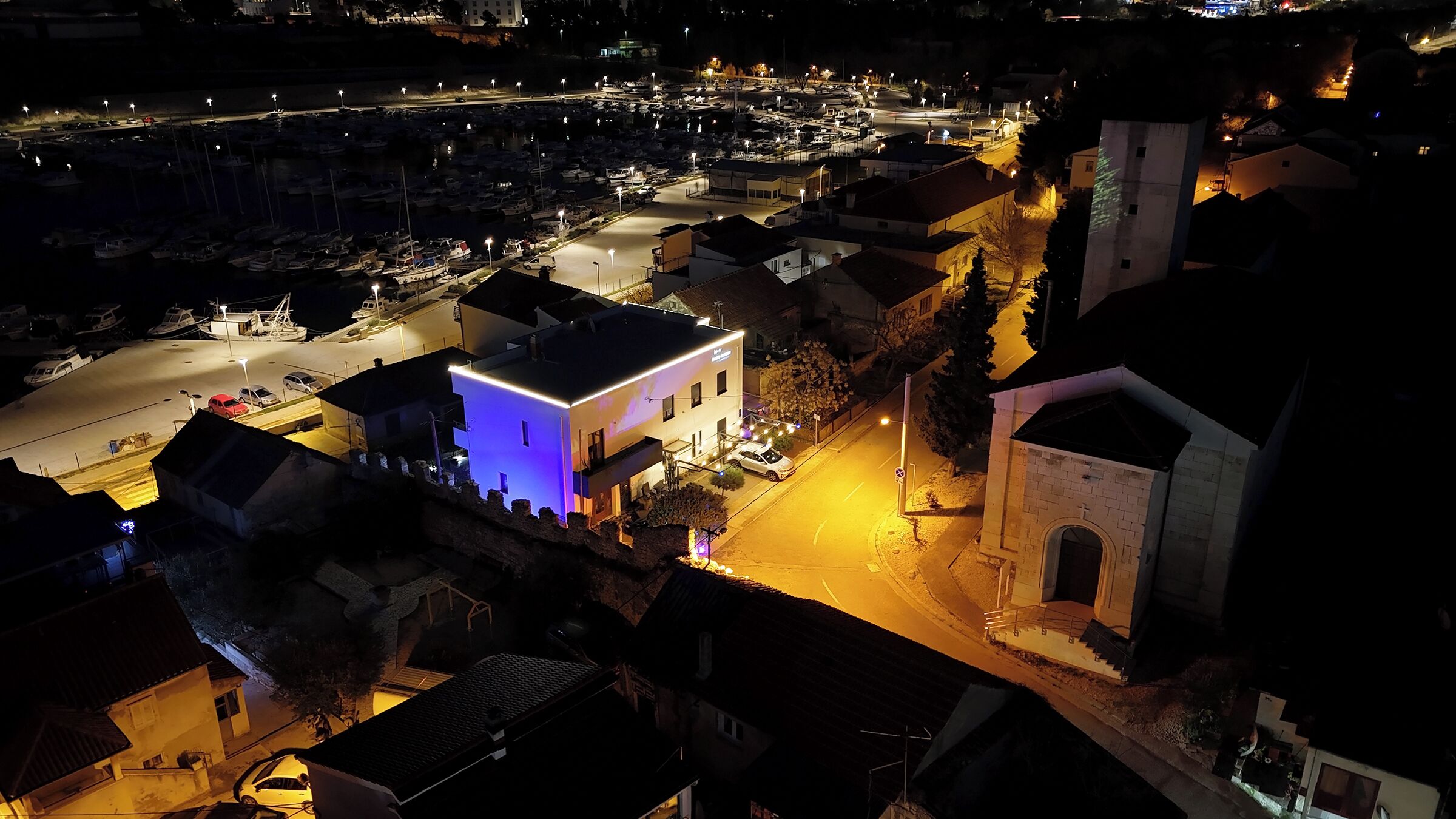 A nighttime view of a marina with boats, a lit building with a blue-lit entrance, and a church with a cross.