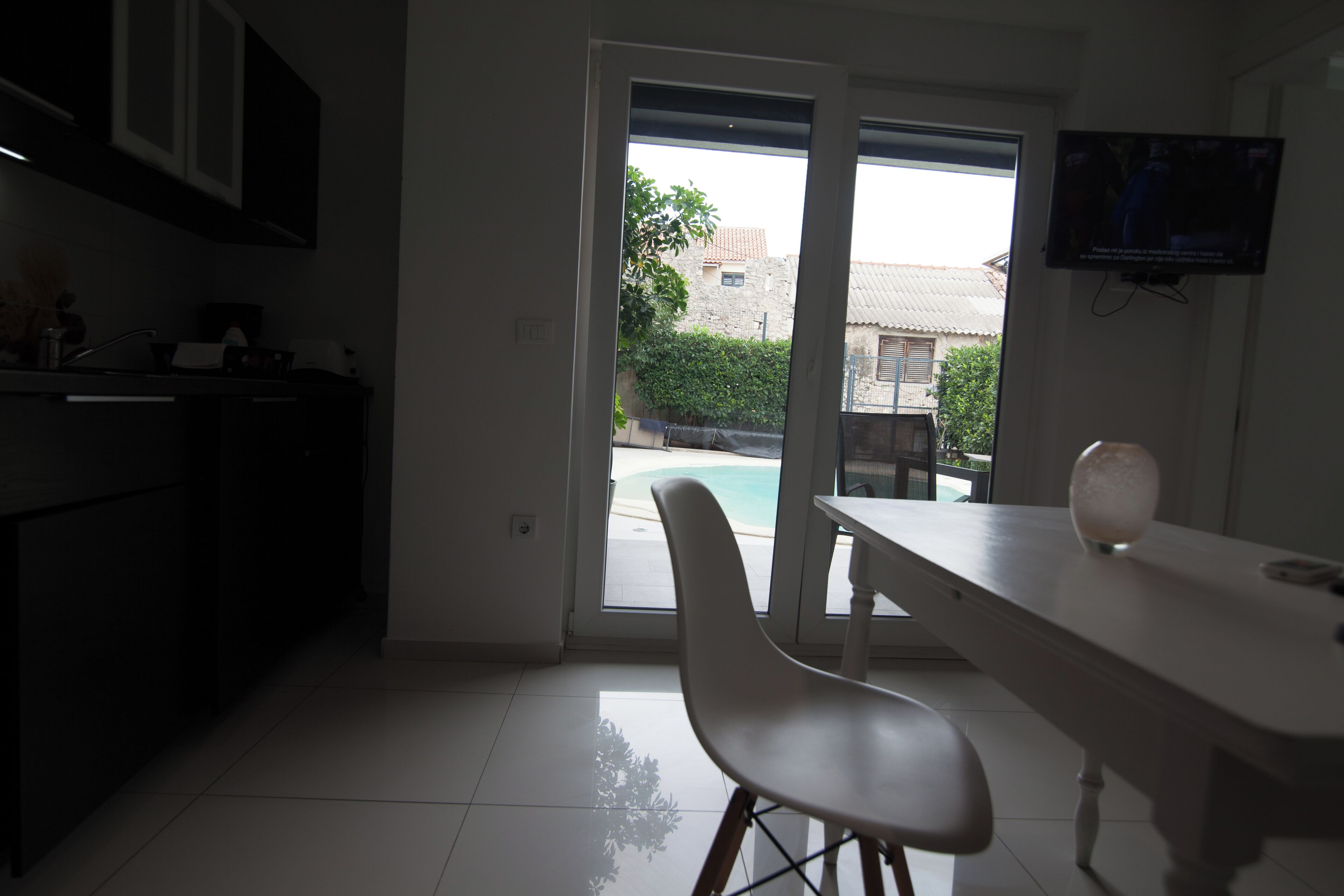 A modern kitchen with a dining area, a TV mounted on the wall, and a view of a swimming pool outside.