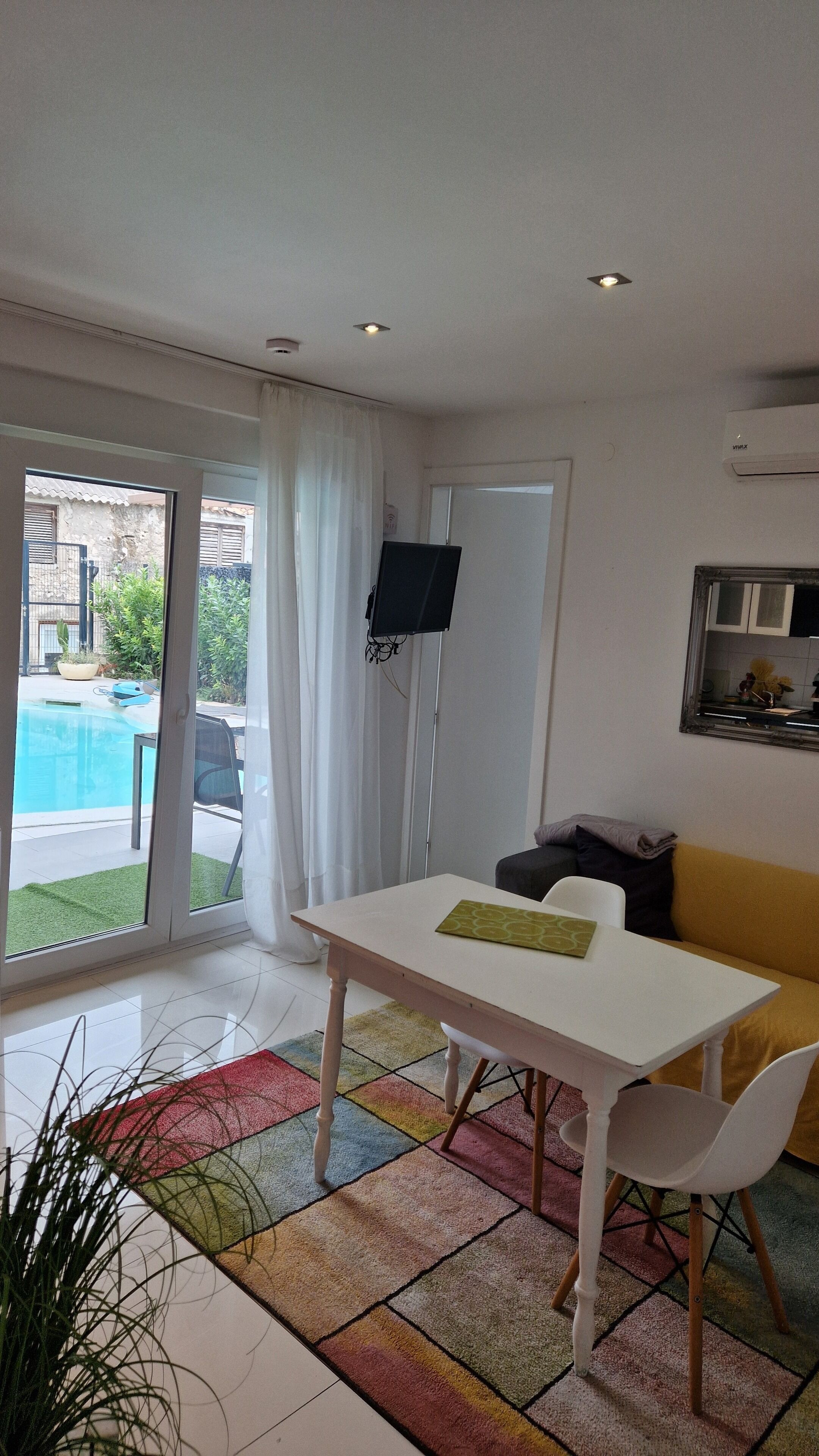 A modern living room with a white table, a yellow sofa, and a large window overlooking a swimming pool.
