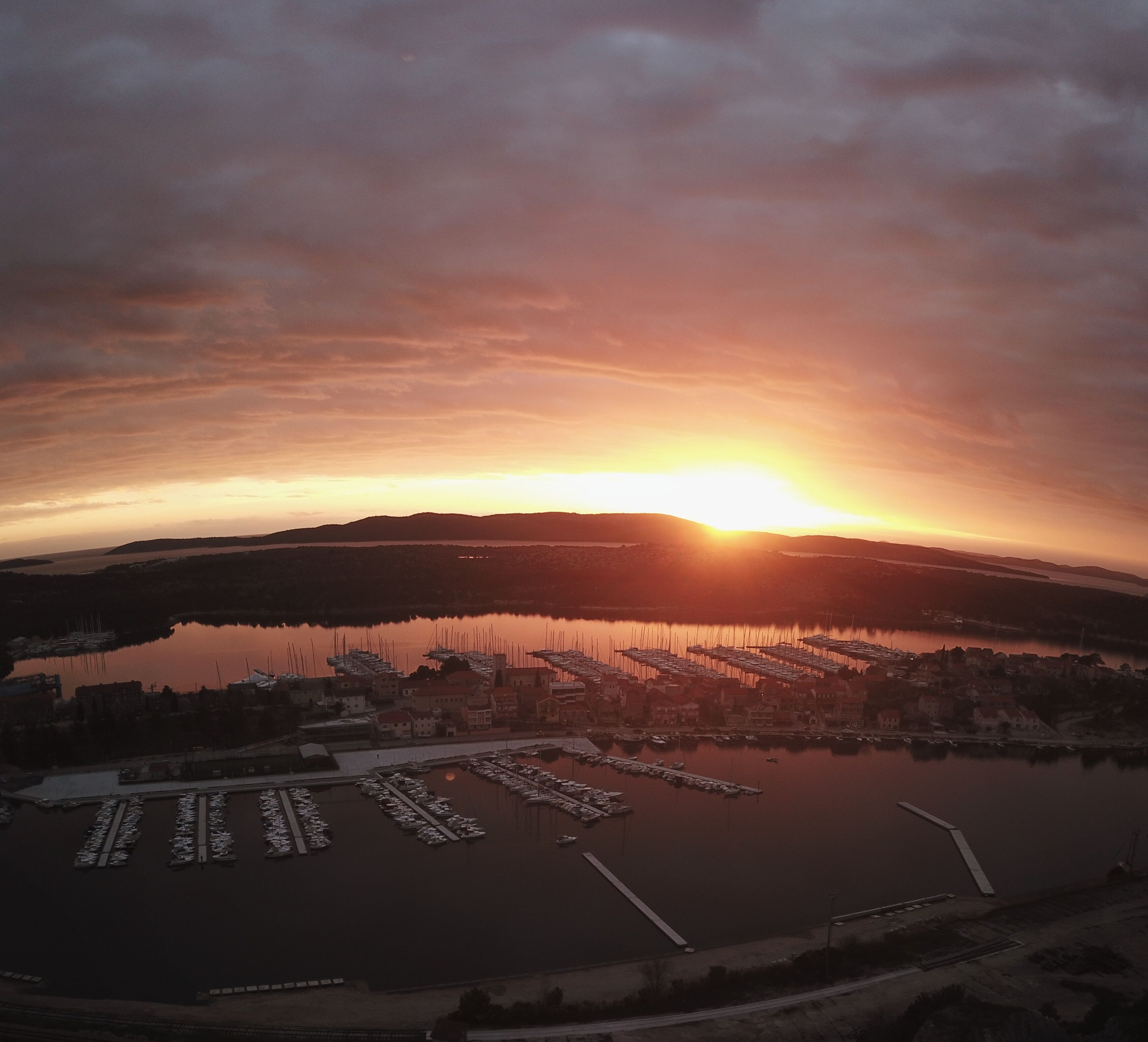 An aerial view of a marina with boats docked, surrounded by hills and a sunset.