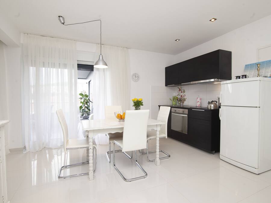 A modern kitchen with white appliances, a dining table with chairs, and a window with sheer curtains.