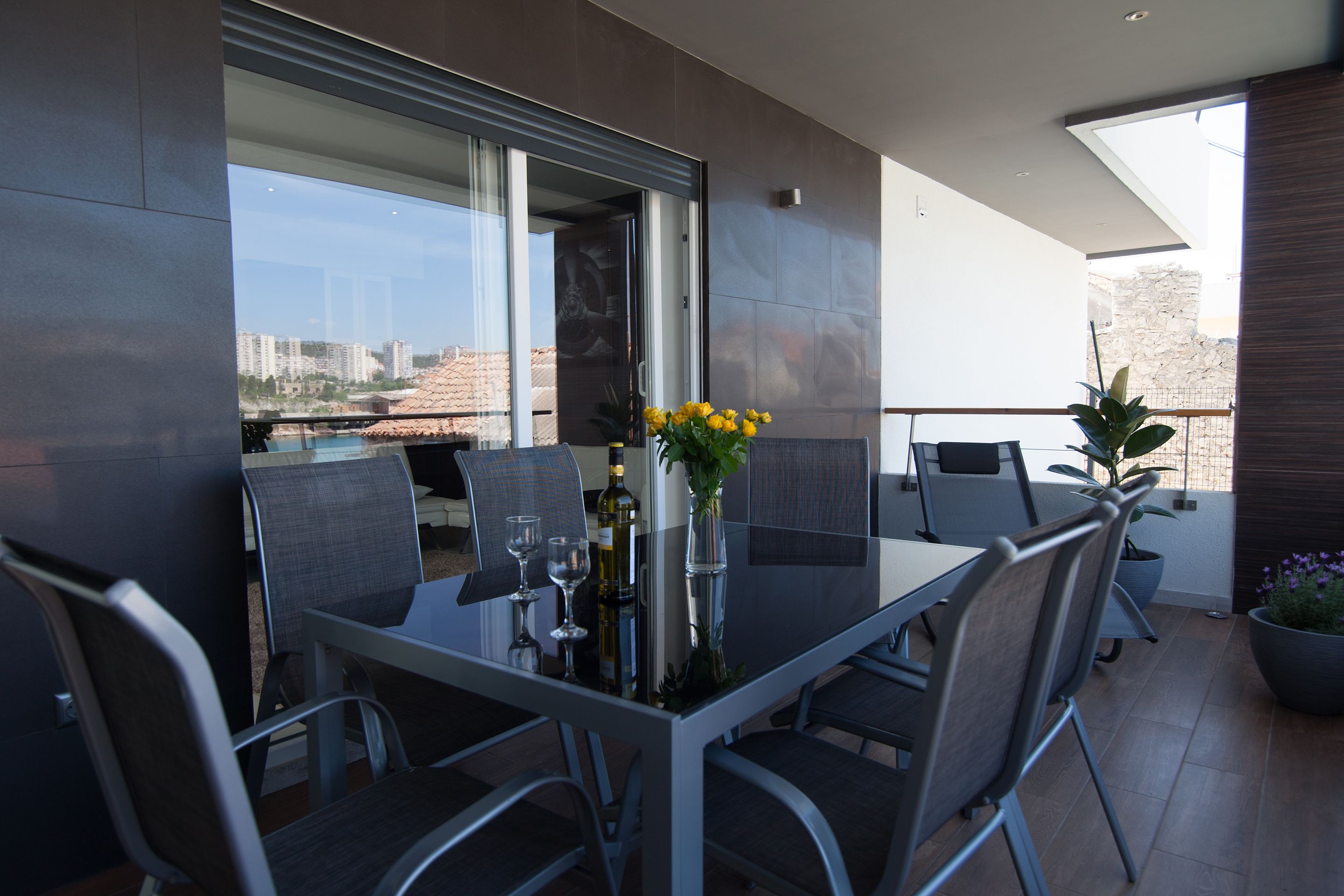A modern outdoor dining area with a glass table, metal chairs, and potted plants.