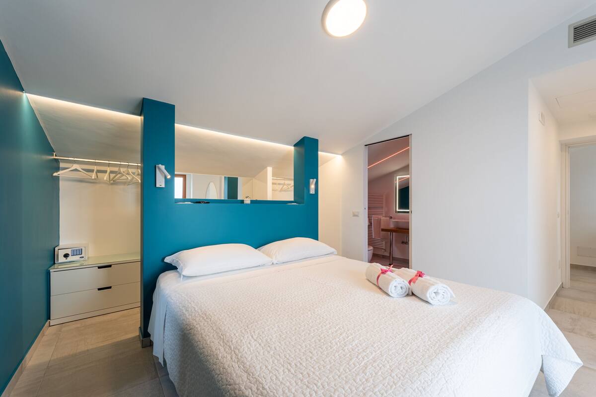 Ein modernes Hotelzimmer mit einem großen Bett, einem eingebauten Kleiderschrank und einem durchgehenden Türrahmen einsehbaren Badezimmer.