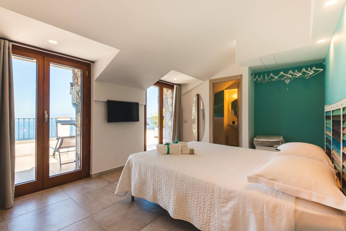 Ein Hotelzimmer mit einem großen Bett, einem Balkon mit Tisch und Stühlen, einem an der Wand befestigten Fernseher und Blick aufs Meer.