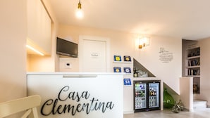 Un hall d’hôtel moderne avec un comptoir de réception portant l’inscription « Casa Clementina », un panneau indiquant les toilettes, un écran numérique, un réfrigérateur rempli de boissons et une bibliothèque.