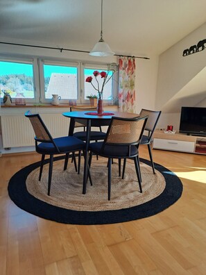 A dining area with a round table, four chairs, a vase with flowers, a hanging light fixture, and a television on a stand. - Apartment Bergblick - Countryside View (Hergatz)