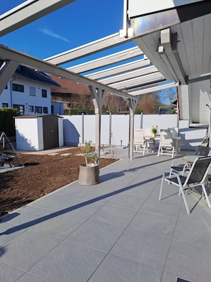 A covered patio area with a seating arrangement, a small garden, and a view of a residential building in the background. - Apartment Bergblick - Countryside View (Hergatz)