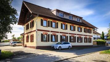 Ein zweistöckiges Gebäude mit schrägem Dach, zahlreichen Fenstern mit Fensterläden und einem geparkten Auto davor.