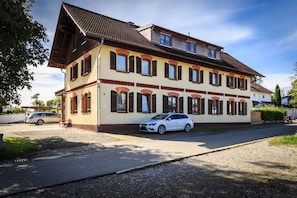 Un bâtiment à deux étages, doté d’un toit en pente, de nombreuses fenêtres avec volets et d’une voiture garée devant.