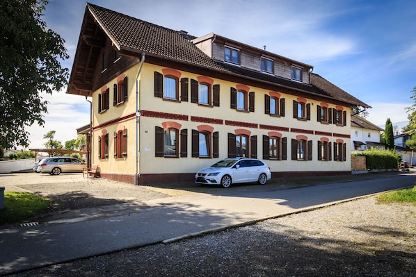 A two-story building with a sloped roof, multiple windows with shutters, and a parked car in front. - Apartment Bergblick - Countryside View (Hergatz)