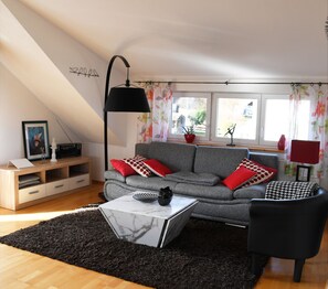 Un salon moderne avec un canapé gris, des coussins à motifs rouges et blancs, un fauteuil noir, une table basse blanche et un lampadaire noir.