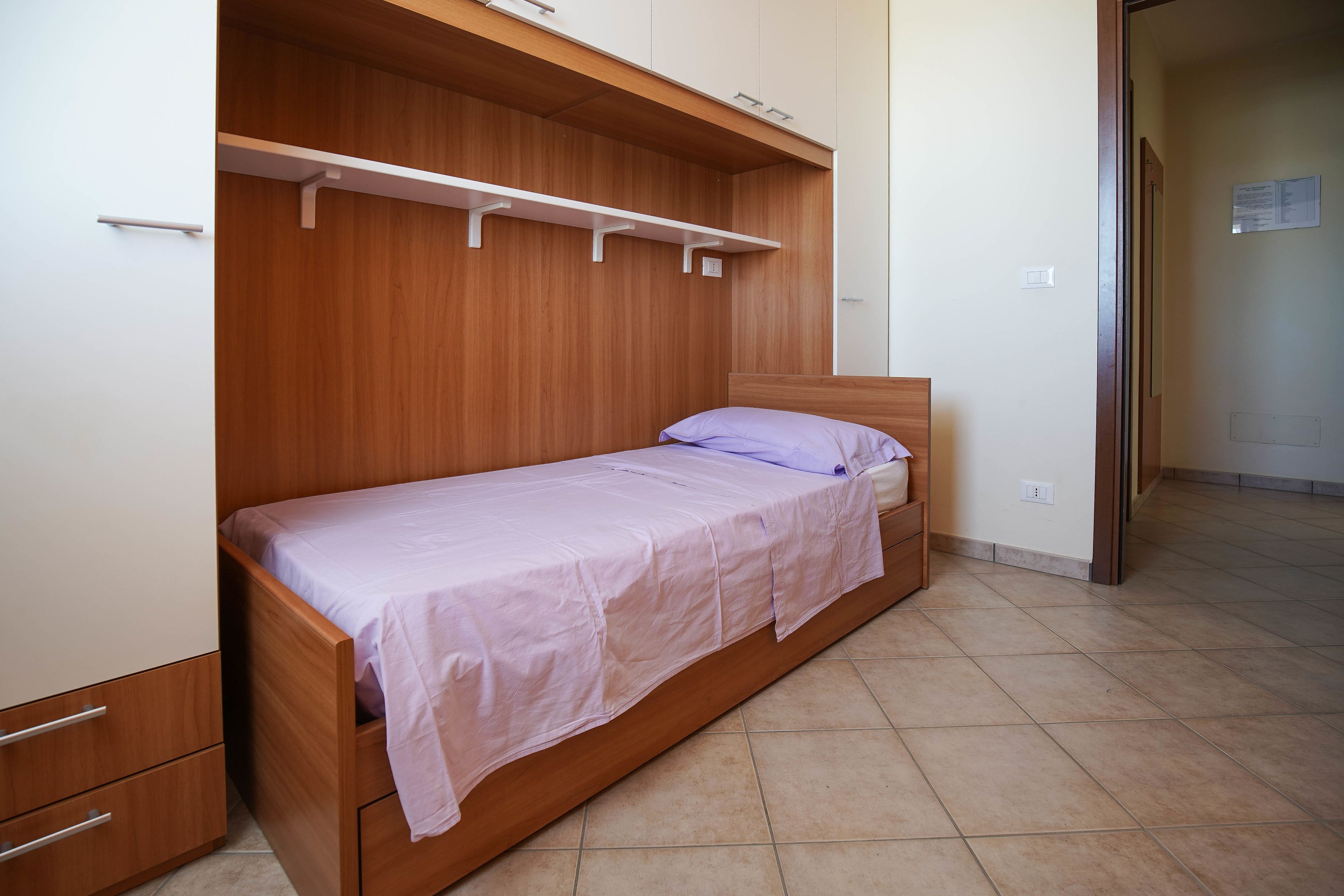 A single bed with a wooden headboard and a white mattress, a wooden wardrobe with a mirror, and a tiled floor.