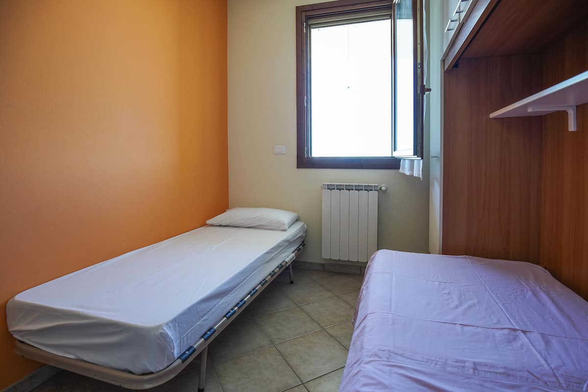 Ein kleines Zimmer mit einem Einzelbett, einem Fenster, einer Heizung und einem Holzschrank.