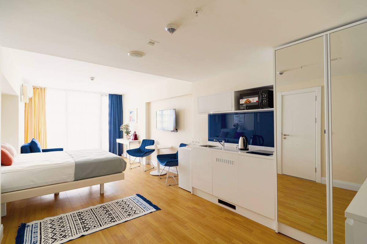 Una habitación de hotel moderna con cama, una kitchenette pequeña, una zona de comedor con sillas y un ventanal con cortinas.