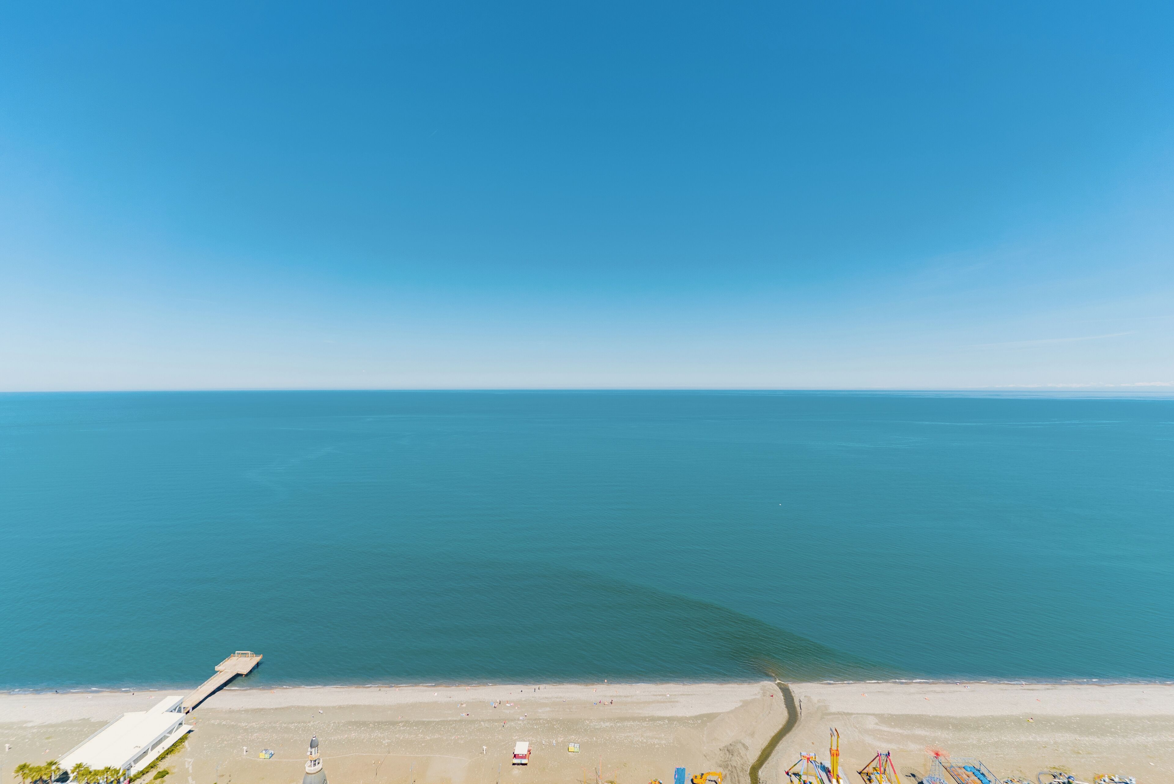 Ovenfra ses en strand med en mole, der strækker sig ud i vandet, en sandet kystlinje og en klar blå himmel.