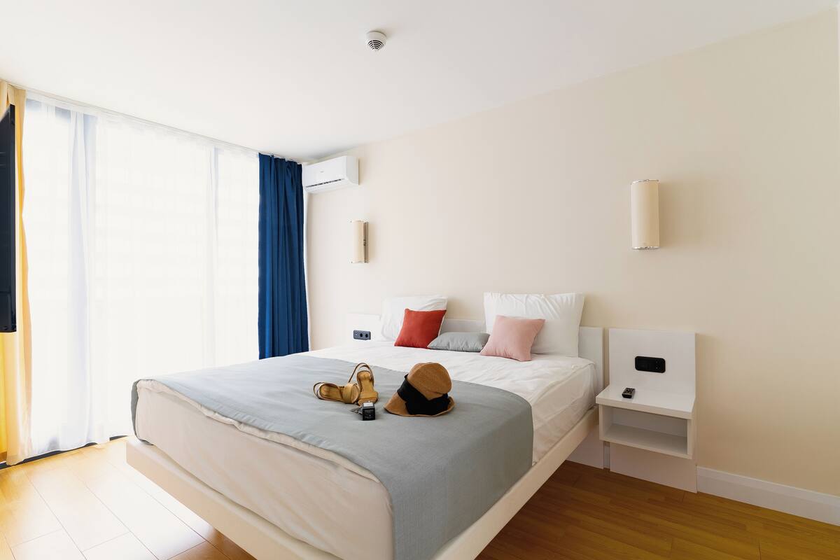 Una cama bien hecha con una colcha gris y blanca, un par de zapatos y un sombrero sobre la cama. Hay una mesita de noche blanca con un teléfono y una pequeña repisa. La habitación tiene un ventanal grande con cortinas azules y un techo blanco con un detector de humo.