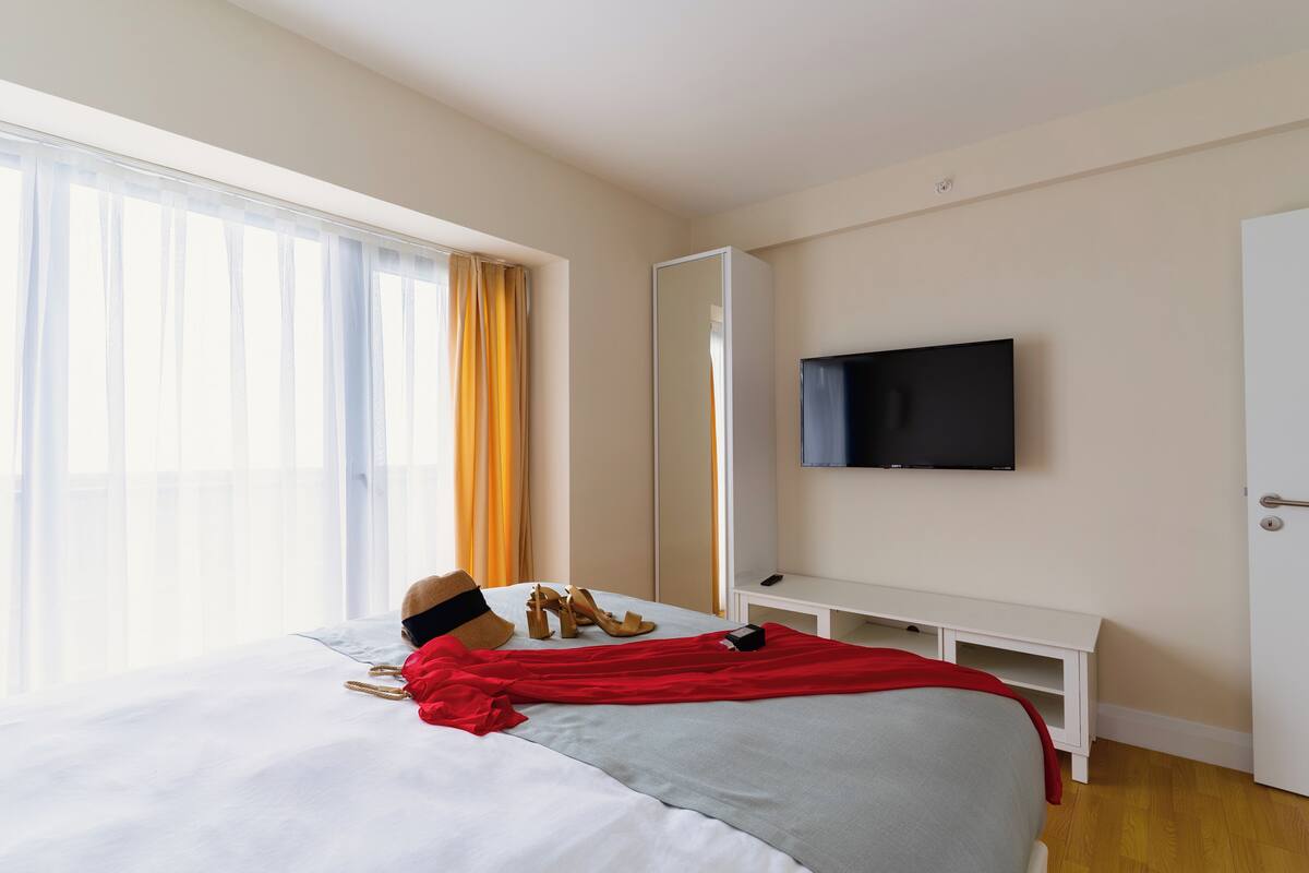 Una habitación de hotel con cama, televisor montado en la pared, espejo y ventana con cortinas.