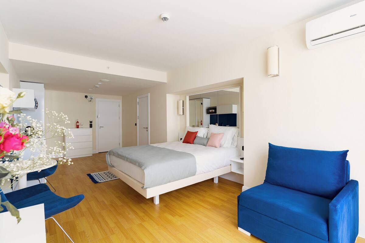 Un dormitorio moderno con una cama, un sillón azul, un espejo y un armario blanco.