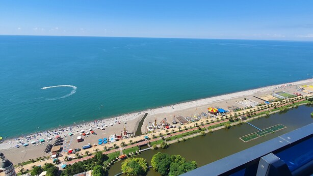 Eine Vogelperspektive zeigt einen Strand, parallel davon fließt ein Fluss, ein Sportplatz und ein kleiner See.
