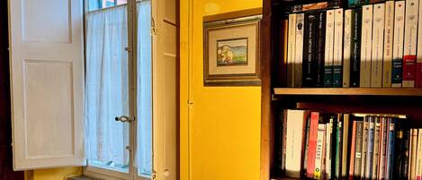 A room with a bookshelf filled with books, a window with curtains, a wall clock, and a table lamp.