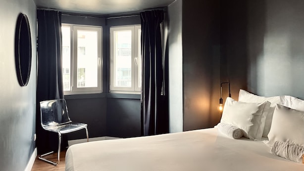 Una habitación de hotel moderna con una cama grande, una silla, dos ventanas con cortinas y una lámpara fijada a la pared.