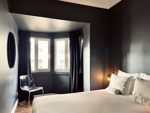 A modern hotel room with a large bed, a chair, two windows with curtains, and a wall-mounted light fixture.