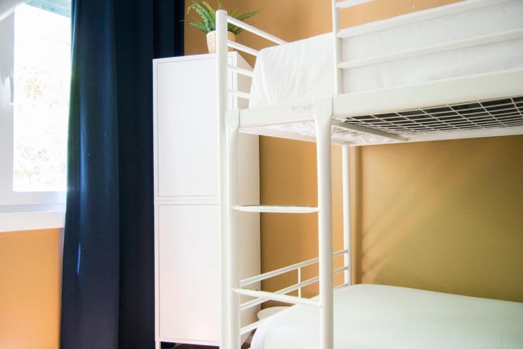 Una cama litera con estructura blanca, una planta pequeña en la parte superior y una ventana con cortinas.