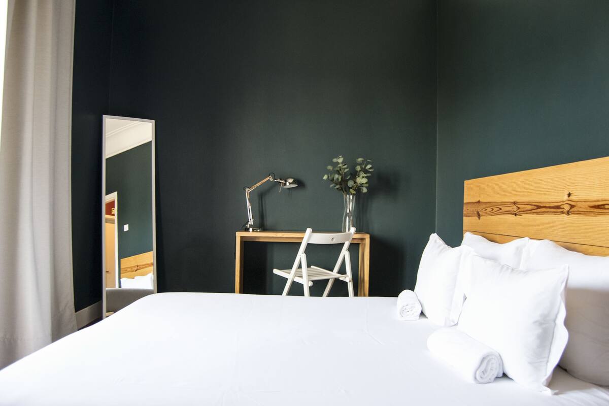 Egy szállodai szoba, amelyben egy nagy ágy, egy fafejű ágykeret, egy éjjeliszemélyke asztallámpával, egy tükör és egy virágos váz található.