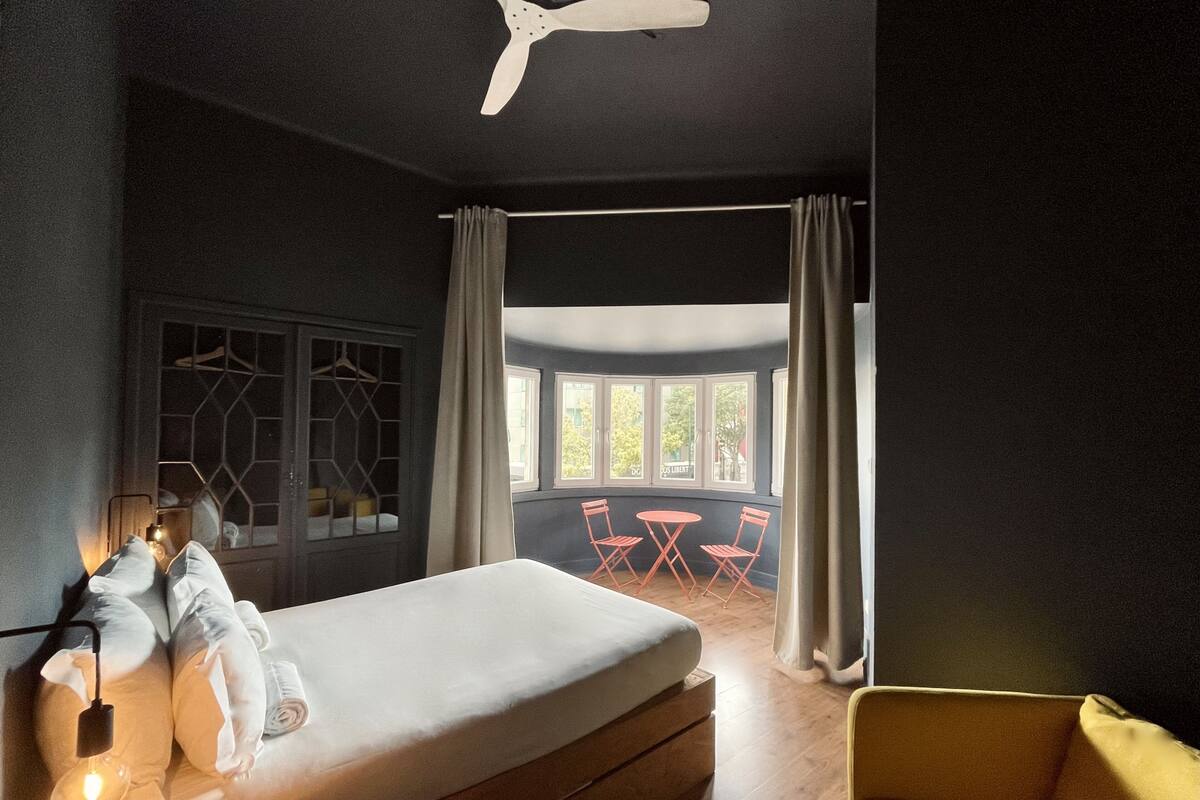 Egy hálószoba, amelyben található egy ágy, éjjeli lámpák, egy mennyezeti ventilátor, egy függönnyel ellátott ablak, és kilátás nyílik egy kültéri ülősarokra.