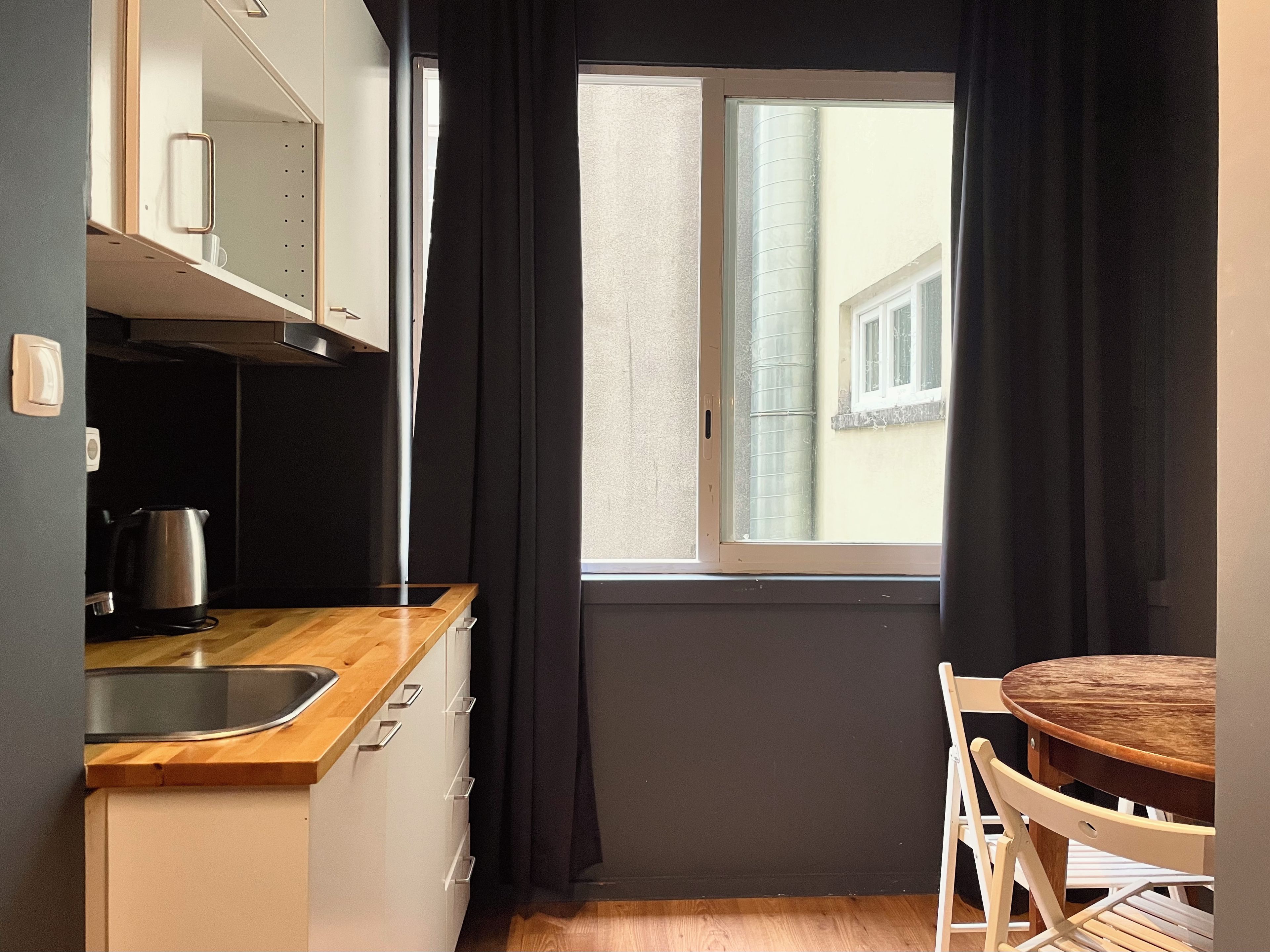 Een kleine keuken met raam, een spoelbak, waterkoker en een houten tafel met stoelen.